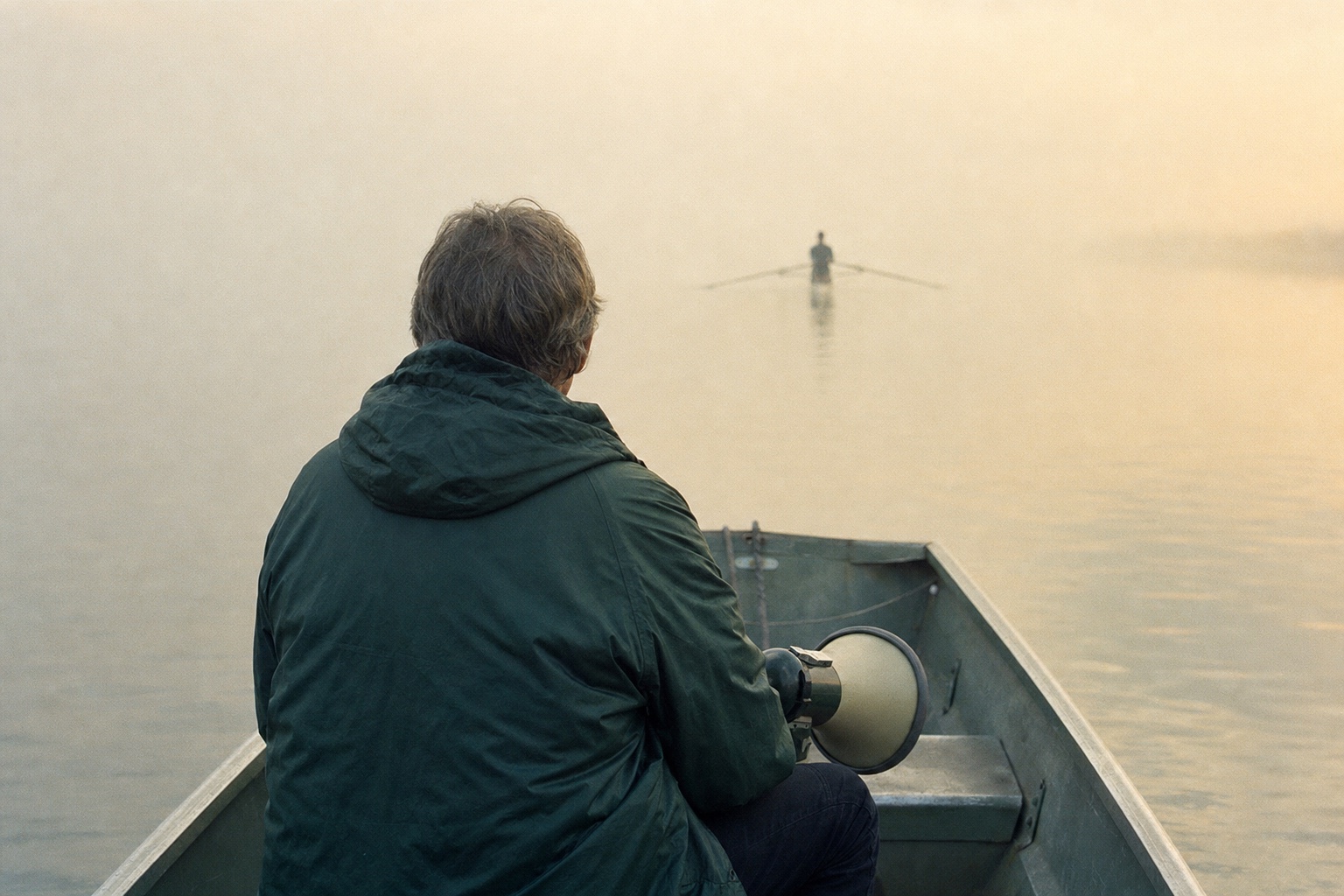From behind: a coach in a dark green jacket sits in a small launch on a misty river, a megaphone on one knee, a single sculler far ahead.